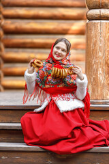 Fototapeta na wymiar A beautiful Russian girl in red traditional Russian clothes holds bagels in her hands in the spring near a wooden house. Festive farewell to winter.