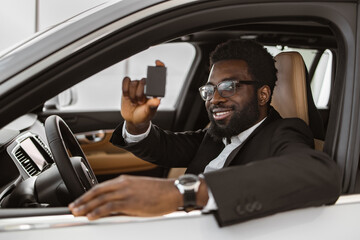 Auto business, car sale, consumerism and people concept. Happy African man taking car key from dealer in bright showroom or salon and showing them to camera.