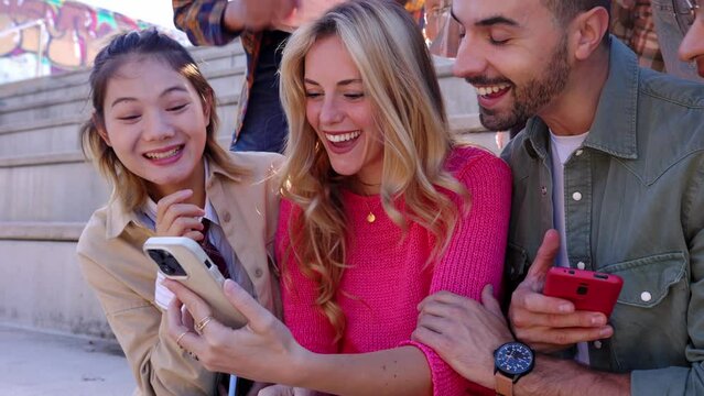 Young multiracial friends having fun looking at mobile phone screen sitting outdoors. Multiracial teenage people laughing together watching funny social media content on smart phone