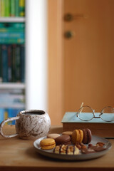 Plate with macarons, cookies, chocolate and nuts, cup of tea or coffee, book and reading glasses on the table. Selective focus. Colorful bookcase in the background.