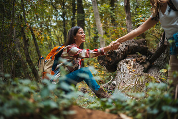 Helping hand - hiker woman getting help on hike smiling happy overcoming obstacle. Tourist backpackers walking in autumn forest. Young caucasian woman helps her biracial friend up a steep trail  - Powered by Adobe