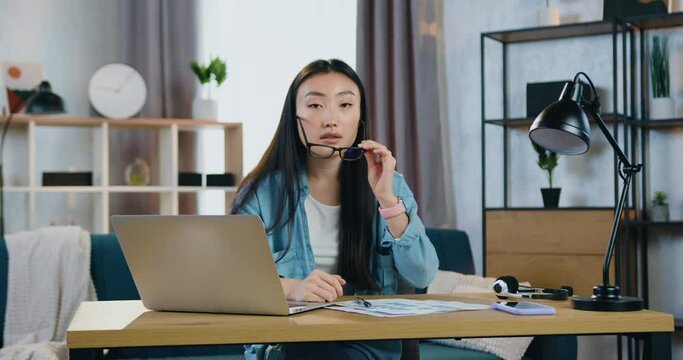 Young asian woman taking off eyeglasses when looking into camera at her workplace in contemporary home office