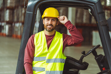 Bearded man, driver, wearing helmet, standing near forklift, looking at camera standing in warehouse