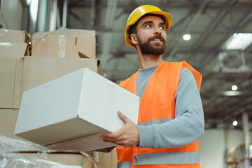 Smiling bearded man worker wearing safety helmet and work uniform holding box, carrying it, mockup