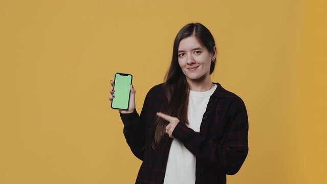 Young attractive woman showing a smartphone with a mockup green screen and cheerfully pointing at it with a finger on a yellow background in the studio. Concept of advice and promotion