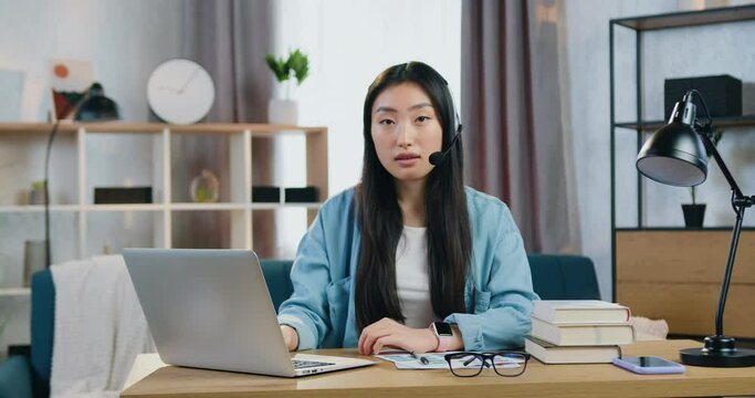 Portrait of young asian woman in headphones with mic looking at camera during video meeting with mentor or tutor. Remote learning concept
