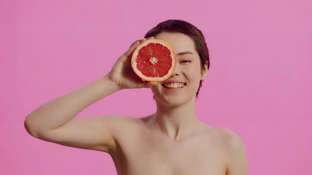 Young attractive woman with clean skin, covering her own eye with a half of the grapefruit and smiling on the pink background. Concept of skin care for health and beauty
