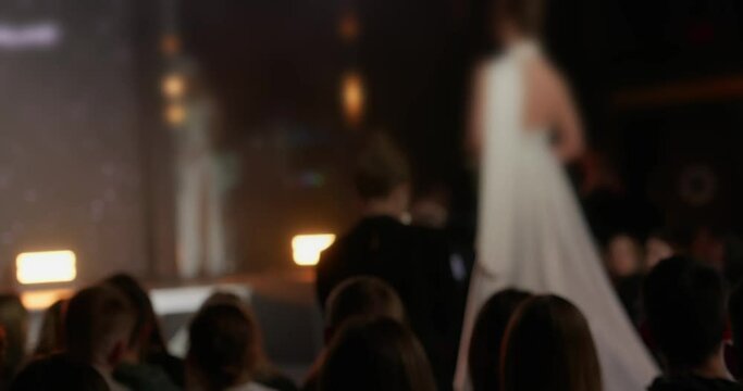 A model in a white dress from the new collection walks down the runway at fashion week towards the stage. The audience is mesmerized by the new dress on the model that passes by them. High quality 4k