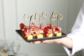 Woman holding plate of tasty canapes with ham and cheese indoors, closeup