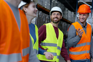 Group of smiling managers wearing hard hats and vests, walking through large warehouse, discussing