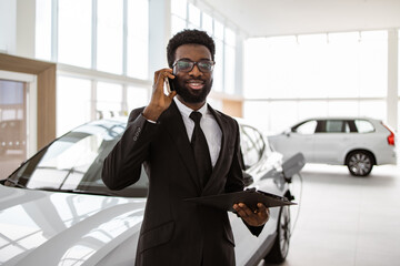 Car dealer holding clipboard in automobile showroom. Smiling salesman using smartphone while having...
