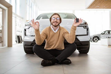 Calm bearded man sitting at lotus pose near new bought car and holding keys in car dealership....