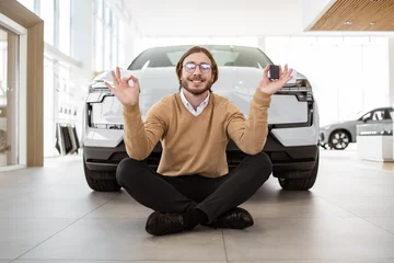 Tuinposter Calm bearded man sitting at lotus pose near new bought car and holding keys in car dealership. Successful buyer in casual outfit rejoices in a new car. Trading concept. © sofiko14