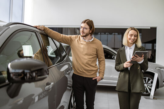 Successful businessman in car showroom, sale of vehicles to customers. Professional female consultant provides all information about new car using tablet to Caucasian male client at car dealership.