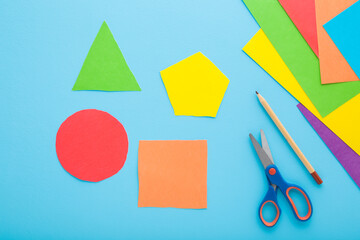 Colorful geometric shapes, scissors, pencil and application paper on light blue table background. Pastel color. Closeup. Child made different forms for learning. Top down view. - obrazy, fototapety, plakaty