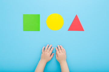 Baby boy hands and cut colorful geometric paper shapes on light blue table background. Pastel...