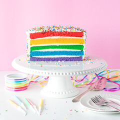 A bright rainbow layered birthday cake surrounded by colourful decorations.