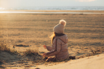 Fototapeta na wymiar A child girl wearing the hat with a pompon sitting on the beach and looking at the sea in sunset time back to the camera