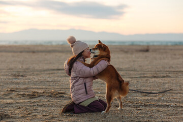 A young girl with a dog in nature. Kid girl hugging a shiba inu dog on the beach at sunset in Greece in winter - 786305194