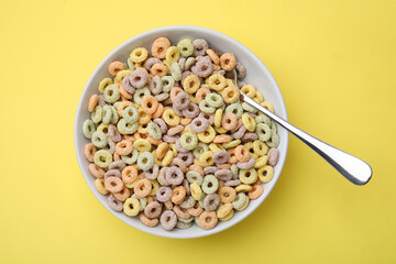 Tasty cereal rings in bowl and spoon on yellow table, top view