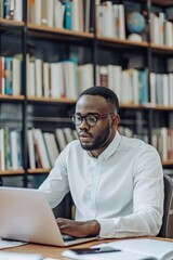 A young African-American man wearing glasses is sitting in a library and using a laptop.