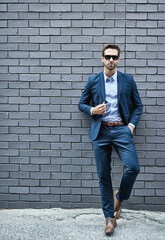 Fashionable, man and wall in suit with sunglasses for fashion, style and trendy in outdoor. Male person, pride and confidence in street for work, corporate or career in mens wear in grey background