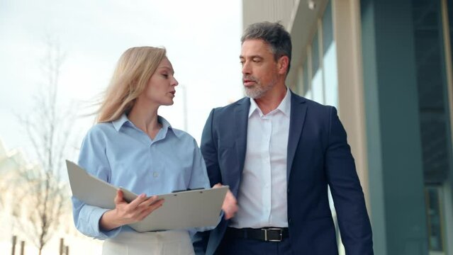 Mature 40s years old man, young adult woman employee walking and discussing project with paper document. Successful middle aged latin hispanic businessman, businesswoman work together on business plan