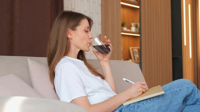 Young caucasian woman sitting on sofa drinking coffee and writing in agenda or journal.