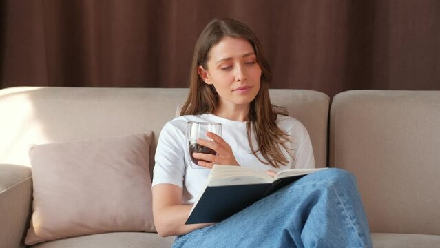 Cheerful caucasian young woman reading a book and drinking cup of coffee and sitting on sofa.