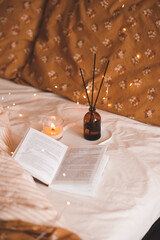 Open paper book with burning candle and diffuser on tray in bed close up. Aromatherapy.