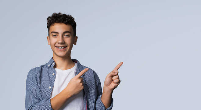 Excited happy black curly haired man in braces, wear denim shirt advertise show peek pointing area for sale slogan text, isolated against grey gray background. Dental care ad advertisement concept.