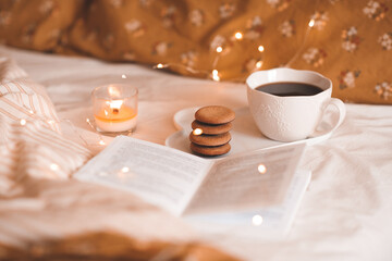 Cup of fresh coffee with biscuits and open paper book in bed over glowing lights close up. Good...