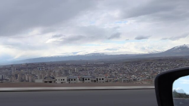 Captivating view from speeding car downhill, showcasing trees, buildings, snow-capped Aragats and Ara mountains on a cloudy day with parallax effect, with partially visible left mirror. Travel concept