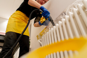 low shot in an apartment cleaner in a yellow apron washes a white radiator with a steam generator