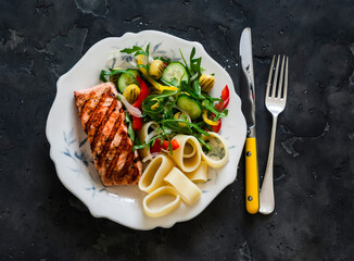 Balanced lunch - grilled salmon, pasta and fresh vegetable salad on a dark background, top view
