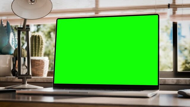 Laptop with a blank screen is placed on a stylish wooden desk within a loft-style interior, with green spaces in the background visible through the window. The camera turns around device