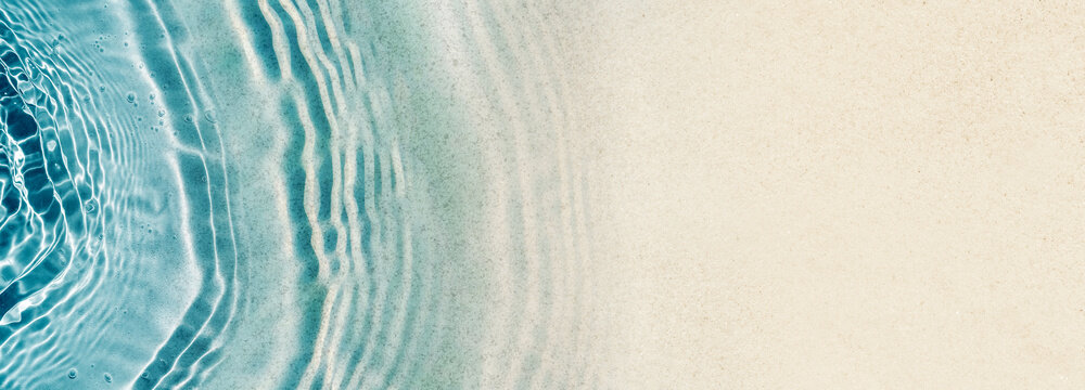 transparent blue water wave on sand beach with sunlight reflections from above, abstract blank beach background banner with copy space for summer holidays, travel and vacation