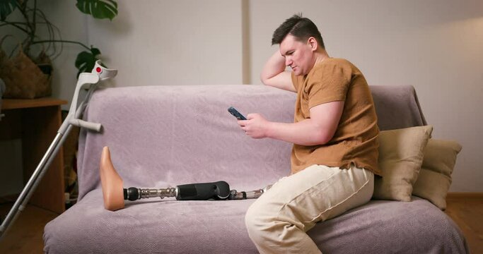 Man amputee with prosthetic leg disability on above knee transfemoral leg prosthesis artificial device using smartphone at home. People with amputation disabilities everyday life