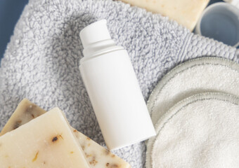 White blank cosmetic bottle on grey folded towel near soap bars in bathroom, mockup