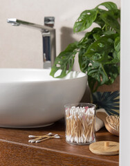 Bamboo cotton swabs near basin and green monstera on wooden countertop in bath, mockup
