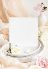 Square card near cream fabric knot and roses on plates close up, copy space, wedding stationery...