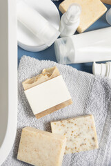 Soap bar with blank label on grey towel near cosmetics in bathroom top view, mockup