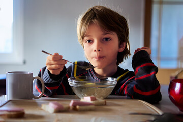 Unhappy male child sits in kitchen table with bowl of cereal in dimly lit kitchen.
