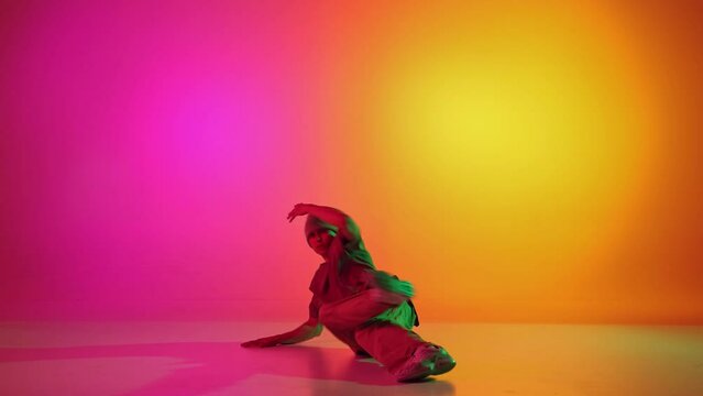 Young athletic man dressed streetstyle clothes dancing on hands in neon light against gradient pink-yellow background. Concept of art, hobby, sport, creativity, fashion and style, action. Ad