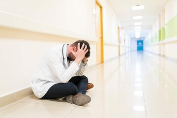 Sad Mid Adult Doctor With Head Hands Sitting Floor Against Wall Corridor Hospital