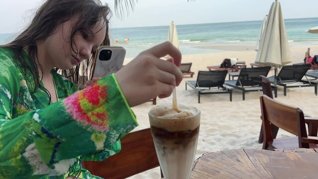 A girl takes a video on her mobile phone, stirring latte Ice latte cappuccino on the beach, a young woman in a green pareo blogs on social networks from the sea, ocean. vacation.