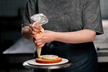 close up female baker in a professional kitchen puts cream from a pastry bag onto a sponge cake