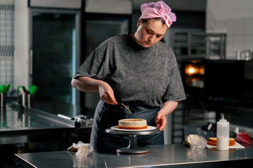 female baker in a professional kitchen distributes cream onto a sponge cake with a spatula - Powered by Adobe
