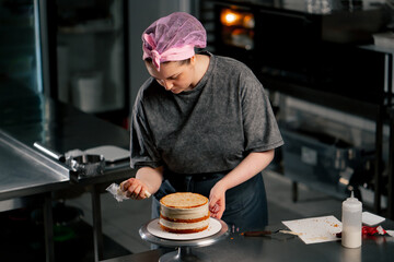 female baker in a professional kitchen puts red cream from a pastry bag onto a sponge cake