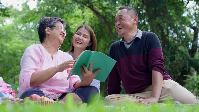 Daughter comes to visit parents and picnic in park, family, leisure and people concept - happy mother, father, elderly caregiver, enjoying outdoor on picnic blanket reading book in park at sunny time